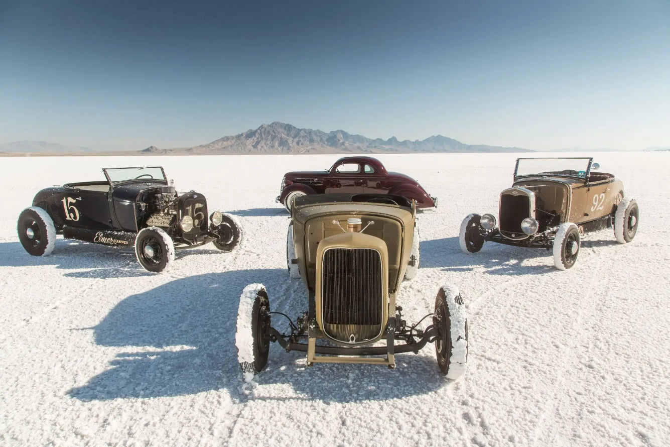 Vintage hot rod cars parked on a white salt flat with mountains in the background.
